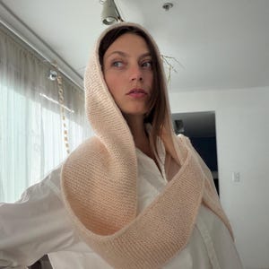 May include: A light peach-colored knitted hood and scarf combination. The hood is designed to frame the face, and the scarf portion drapes around the neck. The soft, neutral color complements the white shirt worn underneath. The knitted texture is visible.