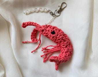 May include: A coral-colored crocheted shrimp keychain featuring a small black eye and dangling yarn legs. Attached to the shrimp is a silver-toned metal lobster clasp and a short string of white beads spelling the name ABINA in black letters.