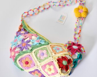 May include: A colorful, handmade crochet bag with a floral design. The bag features a variety of pastel-colored squares and flower appliqués. The strap is made of colorful beads and adorned with crochet flowers. A tag with the text "Knots & Knits" is attached.