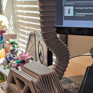 May include: A light gray 3D printed object featuring a rectangular base with triangular cutouts and a tall, flexible, ribbed vertical extension that curves slightly. The structure has a layered texture consistent with additive manufacturing processes.