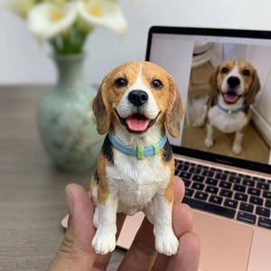 May include: A small, hand-painted beagle figurine featuring a tricolour coat of brown, black, and white, with a light blue collar around its neck. The dog is depicted in a sitting position with its mouth open, showing a pink tongue and a happy expression.