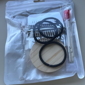 Könnte beinhalten: Ein durchsichtiger Kunststoffbeutel mit einem Stopfwebrahmen aus Metall mit mehreren silbernen Zinken, einer runden hellen Holzscheibe, zwei schwarzen elastischen Haargummis und einem Set langer silberner Nadeln in einer durchsichtigen Röhre.