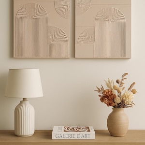 Pode incluir: Dois painéis de arte de parede bege com desenhos esculpidos em arco e círculo. Uma lâmpada branca com base canelada e abajur está em uma mesa de madeira. Um vaso de flores secas e um livro com o texto "GALERIE D'ART" completam a decoração.