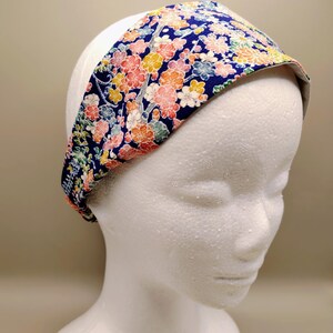 May include: A wide fabric headband featuring a dark blue background with a dense, colourful floral pattern in shades of pink, orange, yellow, and light blue. The headband has a smooth, contoured shape that fits snugly around the head.