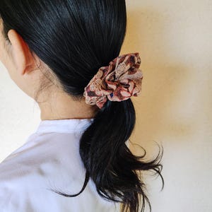 May include: A fabric hair scrunchie featuring a textured, gathered design with a multi-colored abstract pattern in shades of deep red, brown, and muted gold. The accessory is circular, elasticized, and made of a soft, lightweight textile material.