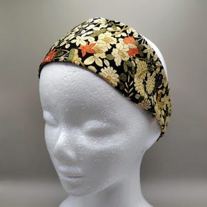 以下が含まれることがあります： 黒い背景にクリーム色、オレンジ色、緑色の花柄が特徴のヘアバンド。 ヘアバンドは幅広で、布製です。 花柄は細かく、バンド全体を覆っています。 白いマネキンの頭に飾られています。