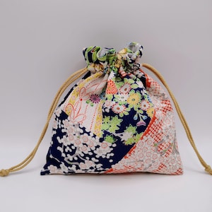 Op de afbeelding: Een buidel met trekkoord voorzien van een levendig patchworkontwerp met bloemenpatronen in marineblauw, wit, oranje en groen. De stof toont ingewikkelde bloesems en geometrische motieven. De tas is bovenaan dichtgetrokken met twee beige koorden.