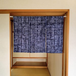 May include: A navy and white patterned curtain hangs in a wooden doorway. The curtain has a textured, woven appearance. The doorway leads into a room with a tatami mat floor and wooden shelves.