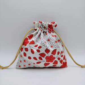 May include: A white drawstring pouch featuring a repeating pattern of red floral motifs and black dotted geometric lines. The bag is cinched at the top with two tan braided cords that extend from the sides, creating a gathered ruffled opening.