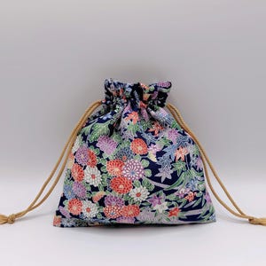May include: A small fabric drawstring pouch featuring a dark blue background with a dense, colorful floral pattern of red, white, purple, and green chrysanthemums and maple leaves. The bag is cinched at the top with two beige twisted rope cords.