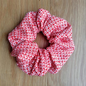 以下が含まれることがあります： 赤と白の模様のシュシュ。生地は、赤い正方形の中に白い正方形が繰り返されるパターンです。シュシュはギャザー入りで円形、髪をまとめるようにデザインされています。アクセサリーは布製です。