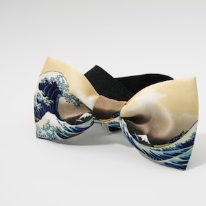 May include: A silk bow tie featuring the iconic "Great Wave off Kanagawa" artwork. The bow tie has a black elastic band and a cream-colored background with blue and brown wave details. A stylish accessory.