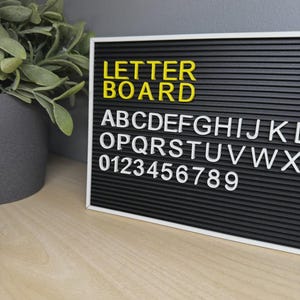 May include: A rectangular black felt letter board with a white frame. The board displays the yellow text "LETTER BOARD" on the top line, followed by white plastic letters "ABCDEFGHIJKL" and "OPQRSTUVWX" on the next lines, and white numbers "0123456789" below.