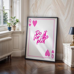 May include: A framed print featuring a playing card design with a pink heart suit and the text "Do it for the plot" in a cursive font. The print is displayed in a room with a window and a small table lamp.