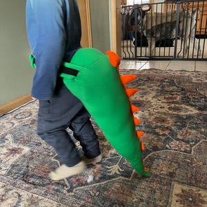 May include: A green plush dinosaur tail with orange spikes is attached to a child's waist. The tail is long and tapered, with a black strap. The child is wearing a blue shirt and dark pants, walking on a patterned rug.