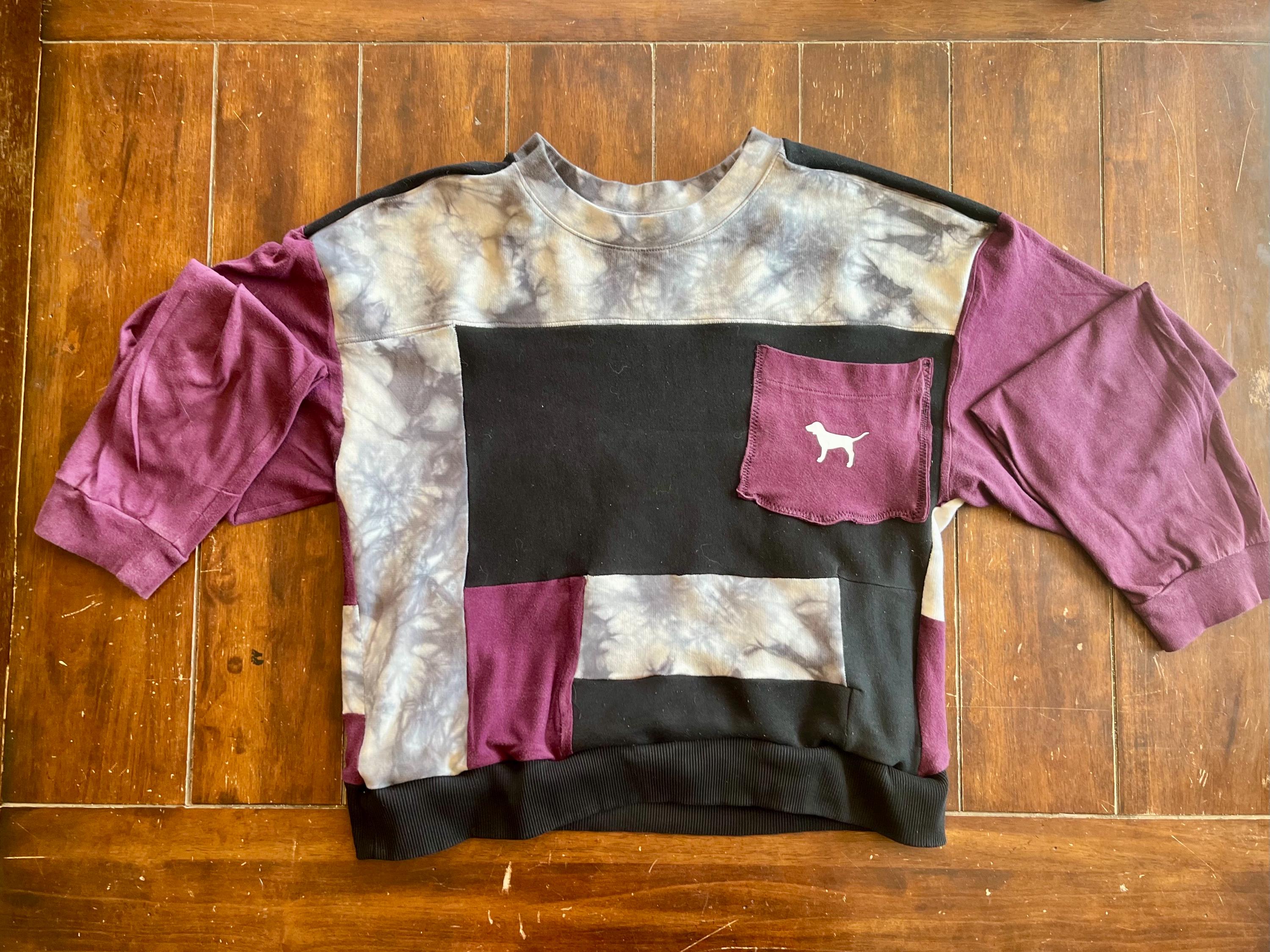 May include: A patchwork sweatshirt with a unique design. The top features a mix of colors, including black, purple, and tie-dye patterns. A pocket on the chest displays a white dog silhouette. The sleeves are a solid purple color.