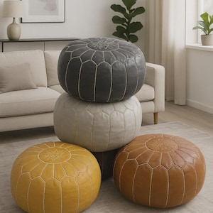 Peut inclure: Quatre poufs ronds en cuir dans un salon. Les poufs sont gris, crème, jaune et marron. Chacun a un motif géométrique cousu et un motif décoratif central. Ils sont empilés sur un tapis neutre. Mesures en cm.