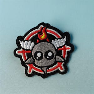 Może przedstawiać: Haftowana naszywka przedstawiająca szarą postać przypominającą czaszkę z dużymi oczami, białymi rogami i małymi ramionami. Projekt zawiera czerwono-biały pierścień, czerwone krzyże i płomień. Czarna obwódka na jasnoniebieskim tle.