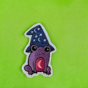 Könnte beinhalten: Gestickter Aufnäher mit einer lila Froschfigur, die einen dunkelblauen Zaubererhut mit weißen Sternen und Monden trägt. Der Frosch hat große schwarze Augen und einen rosa Mond auf dem Bauch. Der Aufnäher hat einen weißen Rand auf einem hellgrünen Hintergrund.