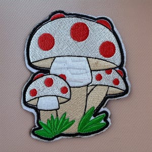 May include: Embroidered patch featuring a cluster of three mushrooms. The mushroom caps are light gray with red polka dots, outlined in black. The stems are tan, and green grass is at the base. The patch has a white border.