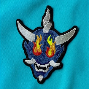 Puede incluir: Parche bordado con un demonio oni azul con cuernos plateados y colmillos blancos. El demonio tiene llamas naranjas y amarillas para los ojos. El parche está delineado en negro, sobre un fondo turquesa.