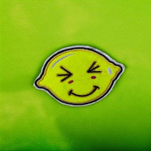 Może przedstawiać: Żółta naszywka w kształcie cytryny z uśmiechniętą twarzą, czarnym obrysem i białym obramowaniem. Cytryna ma zamknięte oczy i dwie małe czerwone kropki na policzkach. Naszywka znajduje się na jasnym zielonym tle.
