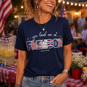 Puede incluir: Una camiseta azul marino de manga corta con el texto blanco 'you had me at' sobre la palabra 'FREEDOM' en un patrón patriótico de bandera estadounidense con estrellas y rayas rojas, blancas y azules, y el texto 'SINCE 1776' impreso debajo.