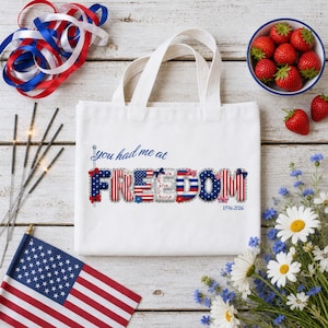 May include: A white canvas tote bag featuring the blue text you had me at and the word FREEDOM in a patriotic red, white, and blue pattern with stars and stripes. The text 1776-2026 is printed in small blue font below the word FREEDOM.