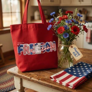 Puede incluir: Un bolso de tela rojo con el texto "SINCE 1776" y la palabra "FREEDOM" en un patrón patriótico de estrellas y rayas. El bolso se muestra junto a un frasco de vidrio lleno de flores rojas y azules y una servilleta plegada con la bandera.