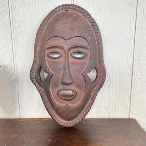 May include: A brown, carved wooden mask with a stylized face. The mask has a prominent nose, open mouth, and almond-shaped eyes. The edges are decorated with a rope-like design, and there is a hole at the top for hanging.