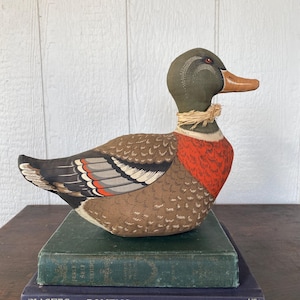 May include: A decorative fabric duck with a brown body, detailed wings, and a red chest. The duck has a green head and a straw collar. It rests on a stack of vintage books, creating a rustic, country aesthetic.