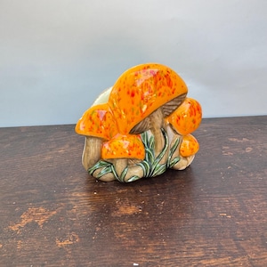 May include: A ceramic napkin holder shaped like a cluster of mushrooms. The mushroom caps are bright orange with red speckles, while the stems are a light tan colour. Green painted grass details are visible at the base of the mushroom stems.