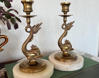 May include: Two brass candle holders, each featuring a dolphin design. The candle holders have a gold finish and are mounted on round, white marble bases. The dolphins have detailed fins and faces. A decorative home accent.