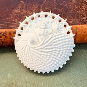 May include: A circular white ceramic ornament featuring a central angel figure surrounded by stars and a sun. The lower portion displays a textured, shell-like pattern with a prominent wave-like curve, while the outer edge has a star-shaped border.