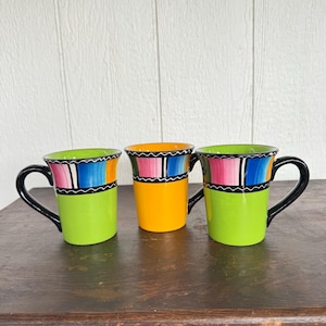 May include: Three ceramic mugs with black handles. Two mugs are lime green, one is orange. Each mug has a colorful band of pink, blue, and orange stripes with a black and white wavy line design. The mugs are on a wooden surface.
