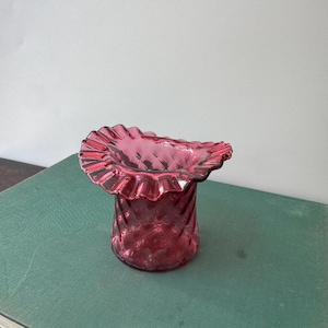 May include: A deep magenta glass vase featuring a wide, ruffled, crimped rim and a textured diamond-patterned body. The vessel has a translucent appearance with a cylindrical shape that flares outward at the top to create a decorative edge.