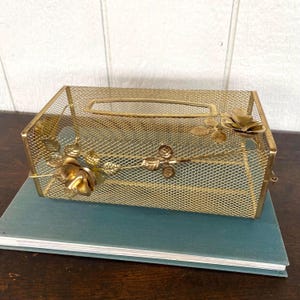 May include: A gold-coloured metal mesh tissue box cover with floral accents. The rectangular box has a rectangular opening on top for tissue dispensing. The box is decorated with gold-coloured roses and leaves.
