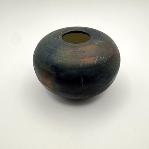 Puede incluir: Un jarrón de cerámica oscuro y redondo con una abertura ancha. El jarrón tiene un acabado moteado con tonos de verde, marrón y negro. La forma general es esférica y la abertura está centrada en la parte superior.