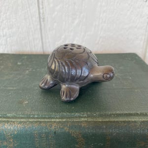 May include: A small, dark grey ceramic turtle figurine with etched details. The turtle has a textured shell with several small holes and is resting on a green surface. The figurine's head and feet are also detailed.
