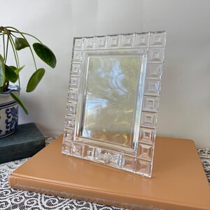 Op de afbeelding: Een rechthoekige fotolijst van helder glas met een decoratieve rand bestaande uit herhalende vierkante, gefacetteerde geometrische patronen. De lijst heeft een gladde, gepolijste afwerking en een centrale opening voor een foto.