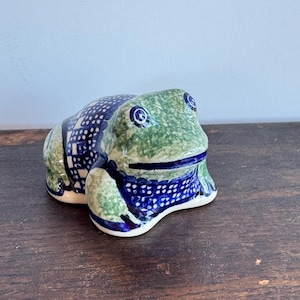 Könnte beinhalten: Keramik-Froschfigur mit grünem und blauem Sprenkelmuster. Der Frosch hat blaue Augen und ein blau-weißes Muster auf dem Rücken. Die Figur steht auf einer Holzoberfläche.