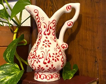 May include: A white ceramic pitcher with a red floral design. The pitcher has a curved handle and a decorative spout. The red design includes flowers and swirls. The pitcher is sitting on a dark red book.