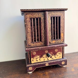 May include: A small, ornate wooden cabinet with a dark burgundy finish and intricate gold-colored detailing. The cabinet features two doors with vertical slats, a drawer with a black knob, and decorative floral and swirl patterns. The cabinet is approximately 8 inches tall.