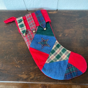 May include: A patchwork Christmas stocking with a red velvet body and various plaid and denim patches. A blue denim patch features a star. The stocking has a red loop for hanging and gold bell accents.
