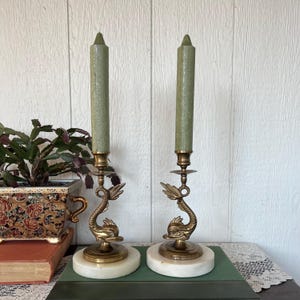 May include: A pair of vintage brass candle holders with dolphin motifs. Each has a white marble base, a brass dolphin body, and a green candle. The candle holders are displayed on a green surface.