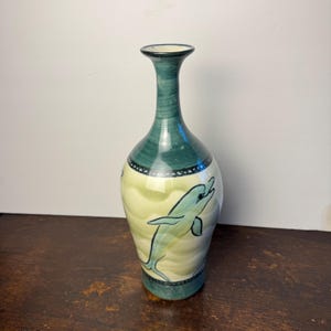 May include: A ceramic vase with a narrow neck and a bulbous body featuring a hand-painted light green dolphin motif against a cream-coloured background with wavy patterns. The top and bottom sections are finished in a deep teal green glaze.
