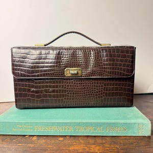 May include: A rectangular dark brown handbag with a textured crocodile skin pattern, featuring a single top handle and a gold-toned metal clasp closure on the front flap. The bag has a structured shape and a smooth, polished finish on the hardware.