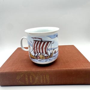 Puede incluir: Una taza de cerámica blanca con asa, con un diseño de barco vikingo con velas a rayas rojas y blancas. La taza está colocada sobre un libro marrón con letras doradas. El diseño incluye elementos de cielo azul y agua.