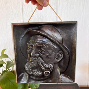 May include: A bronze-colored relief sculpture of an elderly man with a beard, hat, and pipe. The square plaque has a rope for hanging. The man's face is detailed with wrinkles and a stern expression. The sculpture is likely made of metal.
