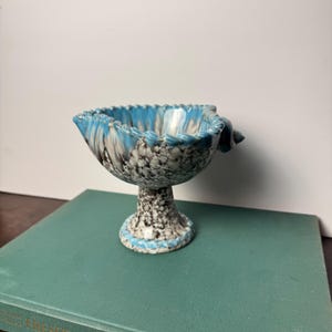 May include: A small pedestal bowl featuring a speckled gray and white pattern on the exterior and base, with a vibrant blue glazed rim. The bowl has a scalloped edge and a small decorative shell-like protrusion on one side of the upper rim.